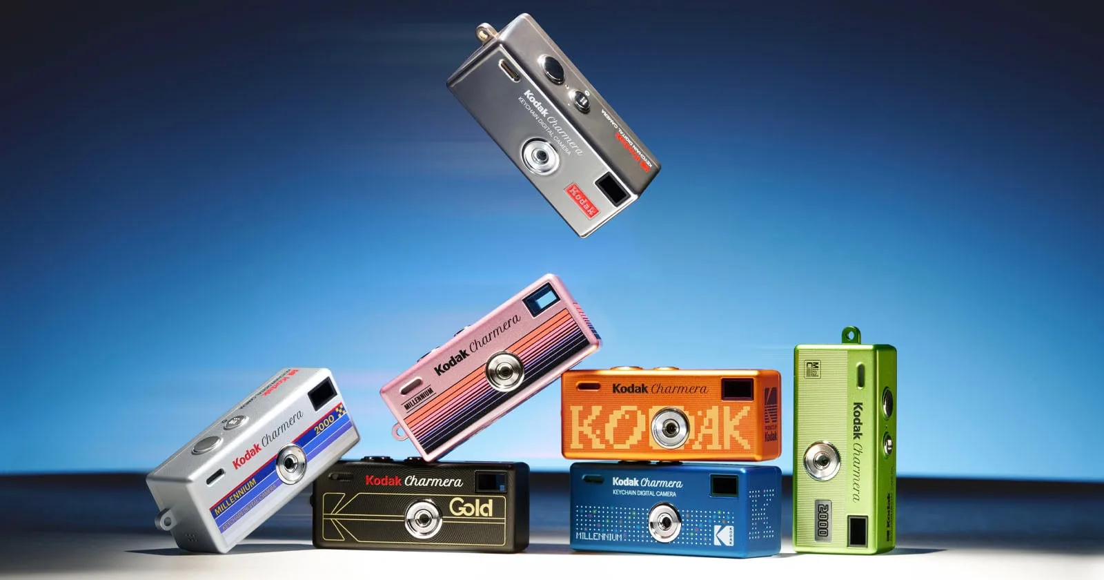 All seven Kodak Charmera Millennium Edition keychain cameras stacked against a blue Y2K backdrop, with the chrome Mirror Silver secret edition floating above them