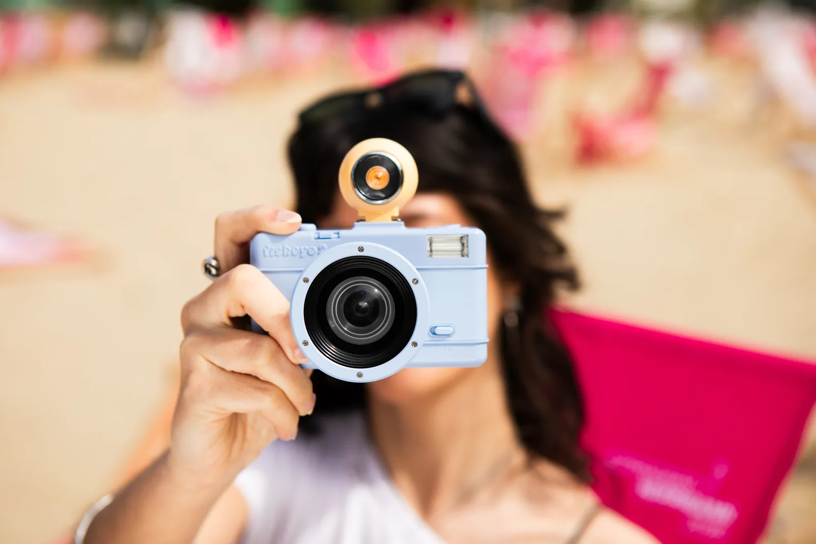 A photographer on a beach holds the periwinkle-blue Lomography Fisheye No. 2 La Mer to her eye, with the round sand-coloured detachable viewfinder mounted on top