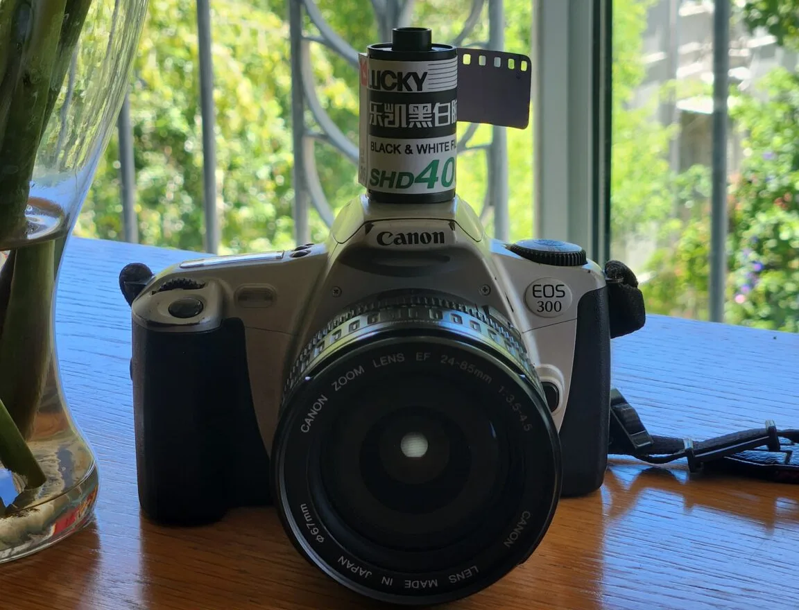 A canister of Lucky SHD400 black and white 35mm film sitting on top of a Canon EOS 300 SLR in front of a sunny window
